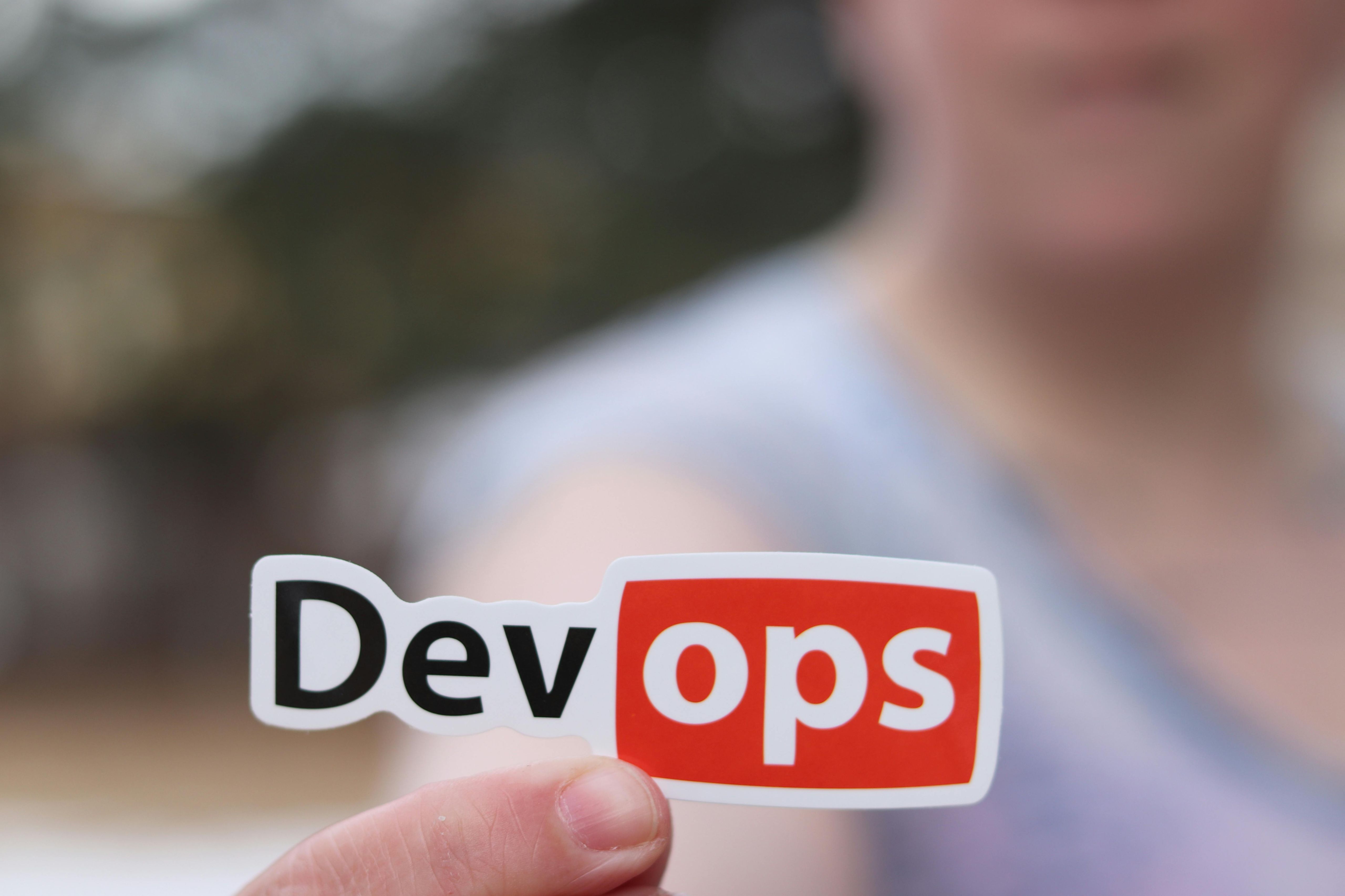 A person holding a sticker with the word "Dev-ops" wright in front of the camera